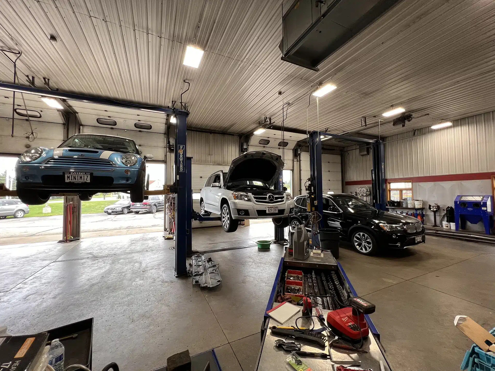 European auto repair services in Sheboygan, WI with NexGen Automotive. The image depicts three cars being lifted: a blue Mini Cooper for wheel alignment, a Mercedes undergoing oil change service, and a black BMW for preventative maintenance. Trust us for expert European auto repair near you.