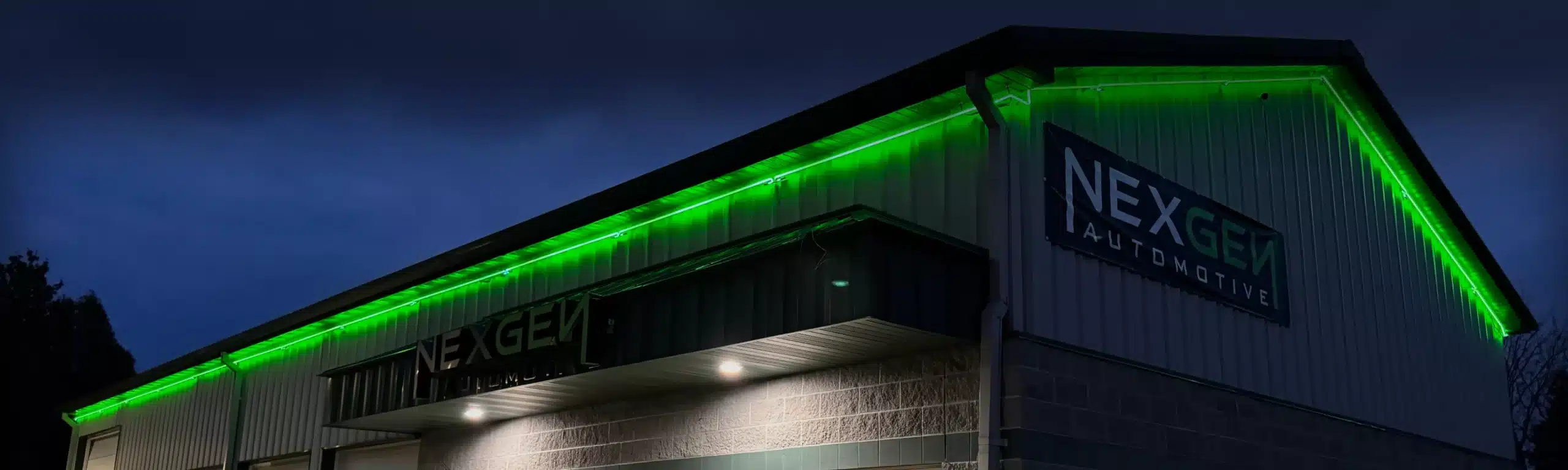 Text: Exterior view of the NexGen Automotive building at night, illuminated by green lights along the roofline.