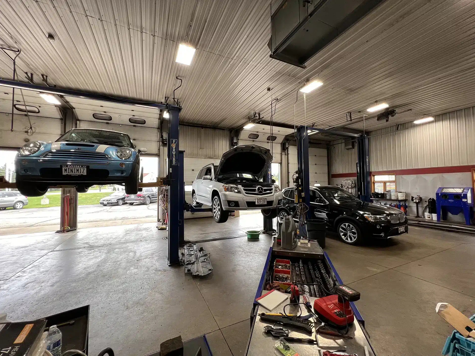 Car maintenance: Should you upgrade or maintain your current vehicle in Sheboygan, WI with NexGen Automotive. Image of 3 black cars, different makes and models all with hoods open and on car lifts for car maintenance at the shop. The middle vehicle has a mechanic underneath performing an oil change.