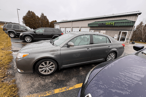 Toyota auto repair shop in Sheboygan, WI with NexGen Automotive. Image of grayish green toyota corolla parked in shop parking lot next to other vehicles that came in for repairs with the NexGen shop and signage in the background.