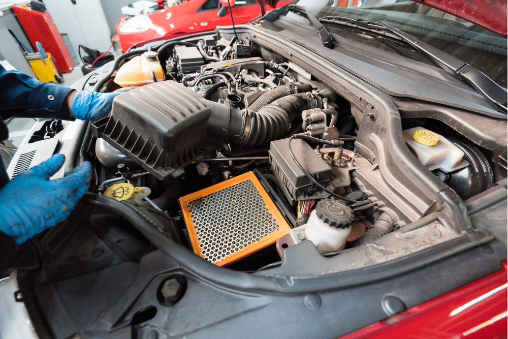 Car maintenance in Sheboygan, WI at NexGen Automotive. Image shows a mechanic wearing blue gloves working under the hood, with the air filter box open and a new engine air filter ready for installation.