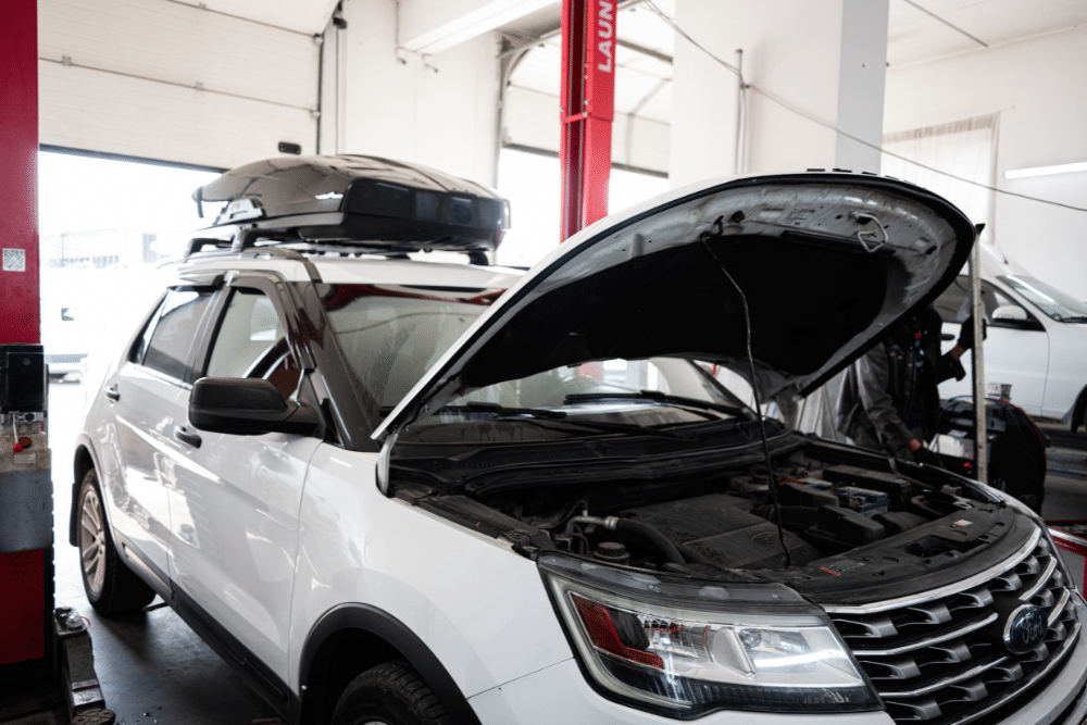 Ford Repair & Maintenance in Sheboygan, WI at NexGen Automotive. A white Ford Explorer SUV is in a mechanic’s shop with its hood open, exposing the engine. The vehicle has a roof cargo box on top. In the background, a red car lift is visible, and there are two people working in the shop.