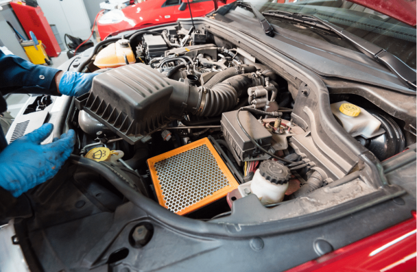 Car maintenance in Sheboygan, WI at NexGen Automotive. Image shows a mechanic wearing blue gloves working under the hood, with the air filter box open and a new engine air filter ready for installation.